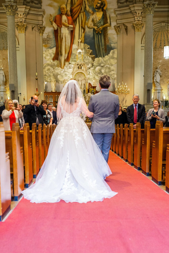 A bride in a flowing white gown and veil walks down the aisle on a red carpet, accompanied by a man in a gray suit