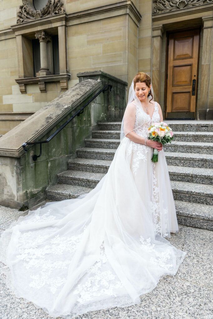 A bride in a lace wedding dress and veil stands on stone steps