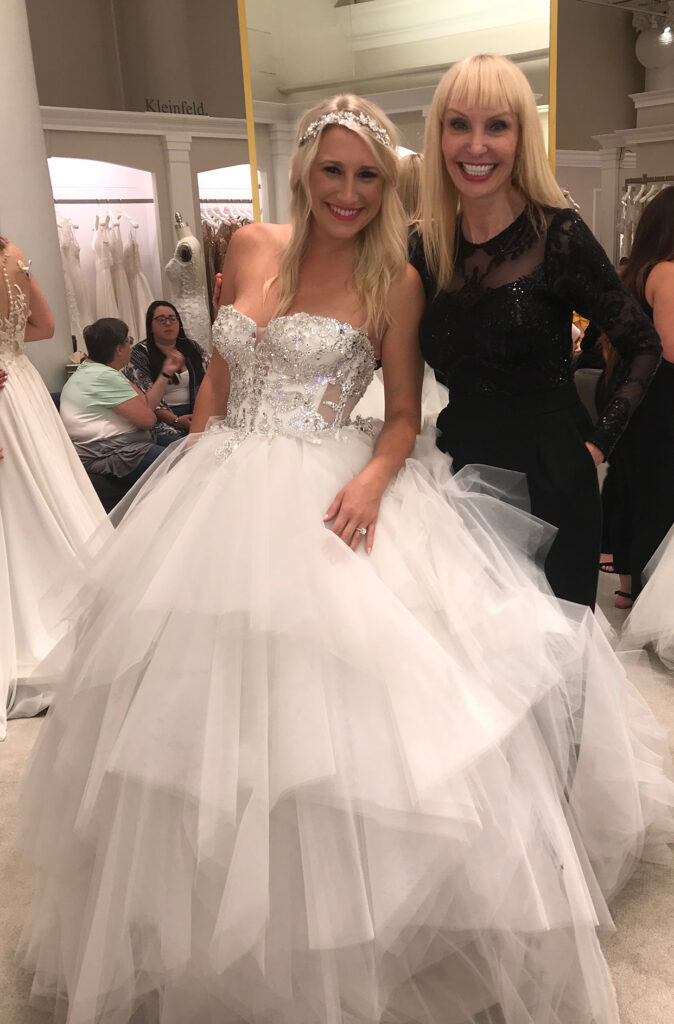 A smiling woman in a sparkling, strapless wedding gown with a voluminous tulle skirt stands next to another woman in a black dress at a bridal boutique