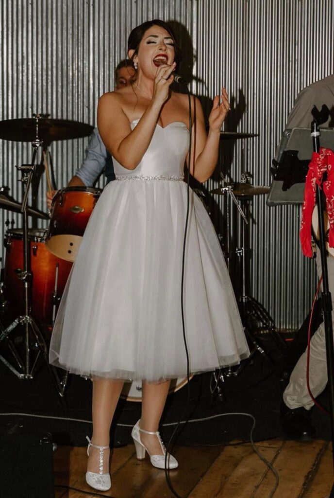 A woman in a white strapless dress passionately sings into a microphone on stage, with a drummer playing in the background