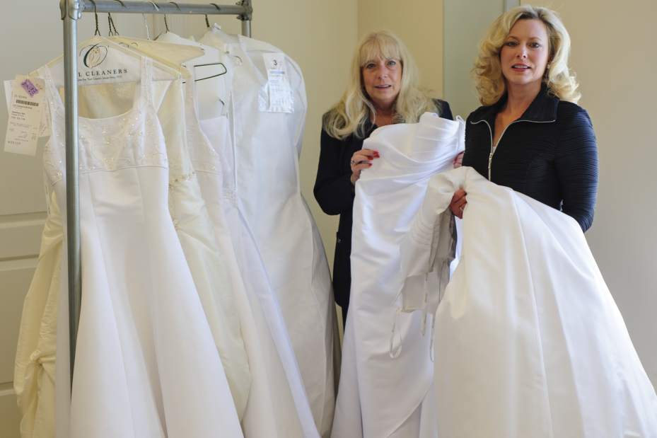 Two women stand beside a rack of white wedding dresses, holding gowns and smiling