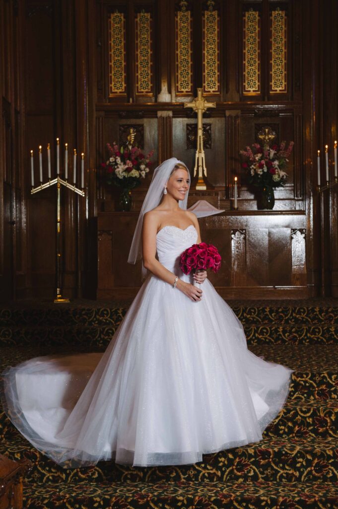 A bride in a strapless white wedding gown stands in a church, holding a bouquet of red roses