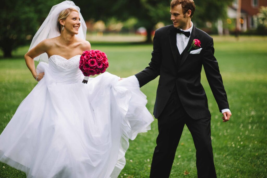 A bride in a white gown holds a vibrant bouquet of pink roses while smiling at a groom in a black tuxedo
