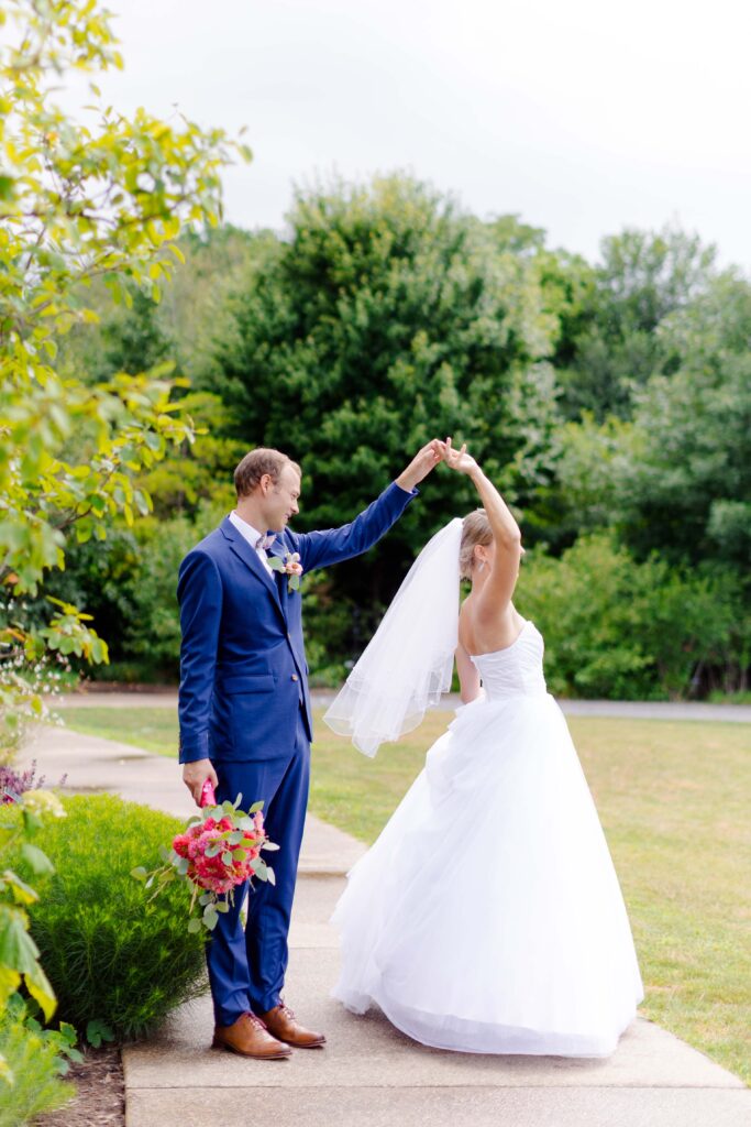 A groom in a blue suit twirls his bride, who is wearing a white wedding dress and veil, on a garden path