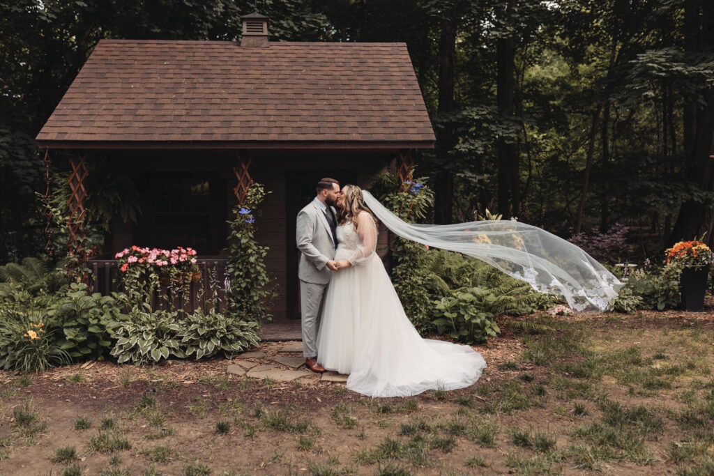 A bride and groom share a kiss in front of a rustic wooden cabin surrounded by lush greenery and colorful flowers
