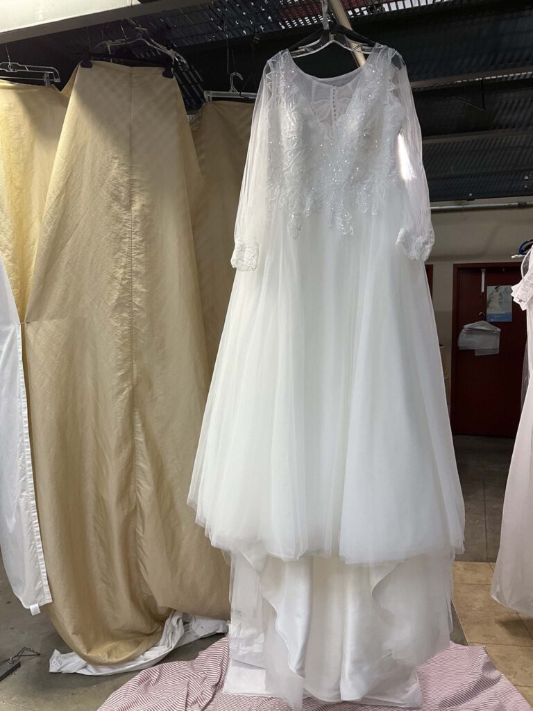 A white wedding dress with lace and bead detailing on the bodice and long sleeves hangs on a rack