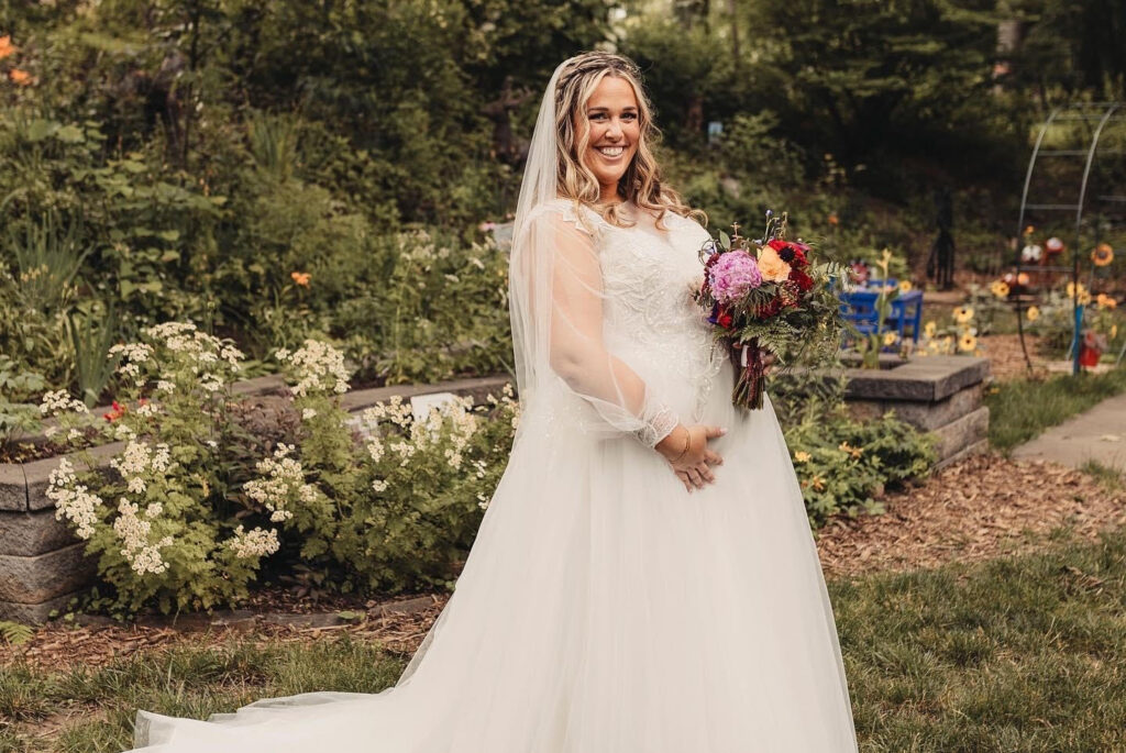 A bride in a flowing white gown and veil smiles while holding a colorful bouquet of flowers