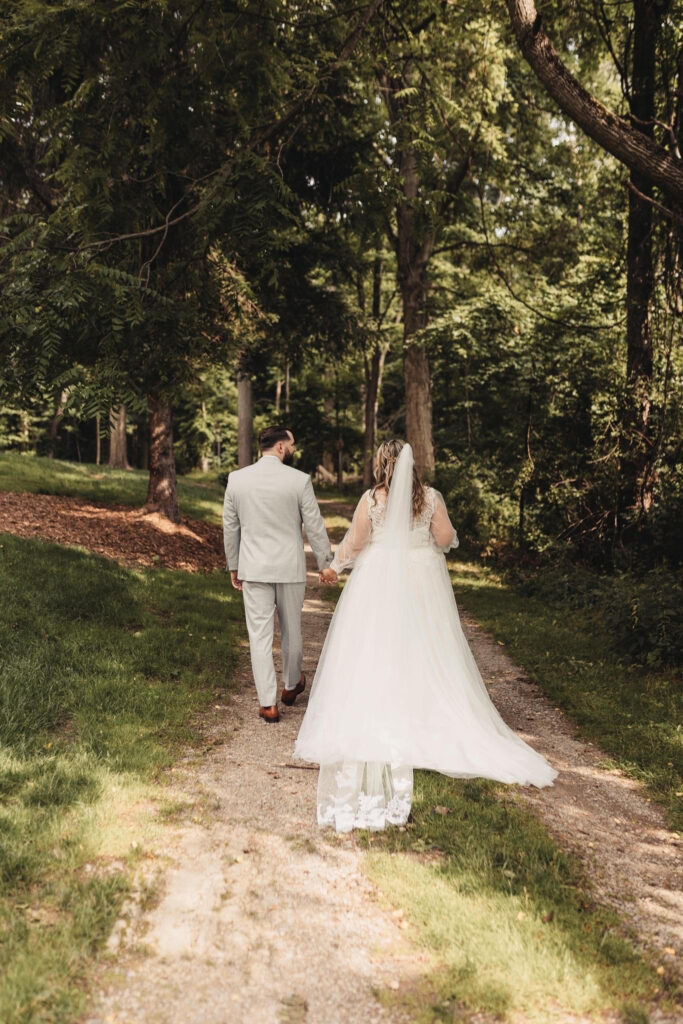 A bride and groom walk hand in hand down a forest path, surrounded by lush greenery