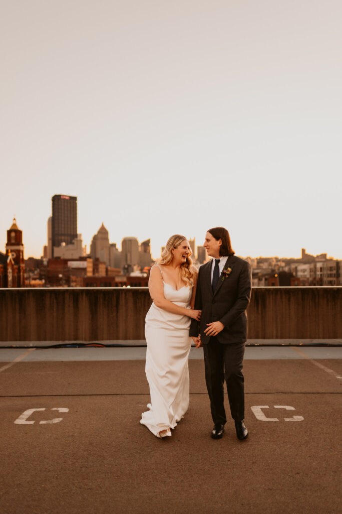 A bride and groom walk hand in hand on a rooftop, smiling and looking at each other