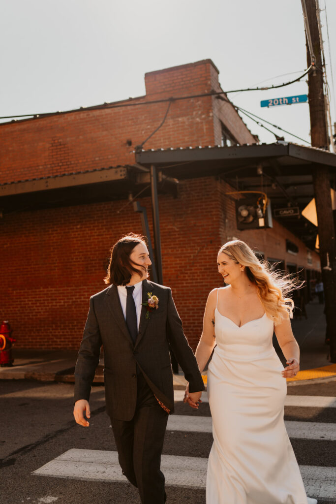 A couple dressed in wedding attire walks hand in hand across a street
