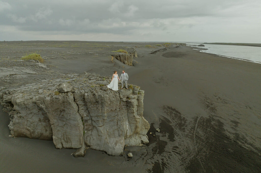 A couple in wedding attire stands on a rugged cliff overlooking a vast, desolate landscape