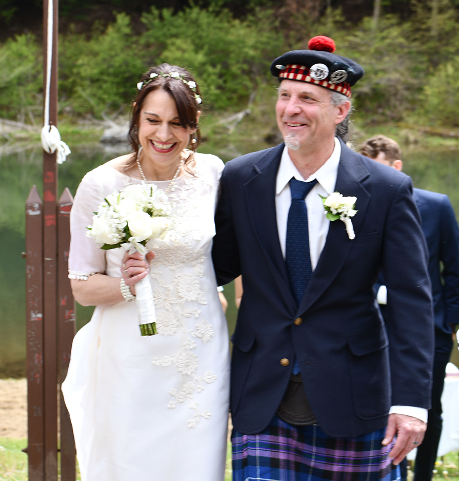 A joyful bride in a white dress holds a bouquet of white flowers, walking arm-in-arm with a man wearing a dark blazer
