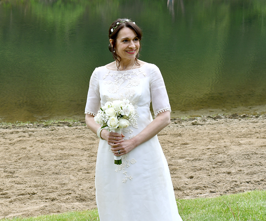 A woman in a white wedding dress stands by a serene lake, holding a bouquet of white roses