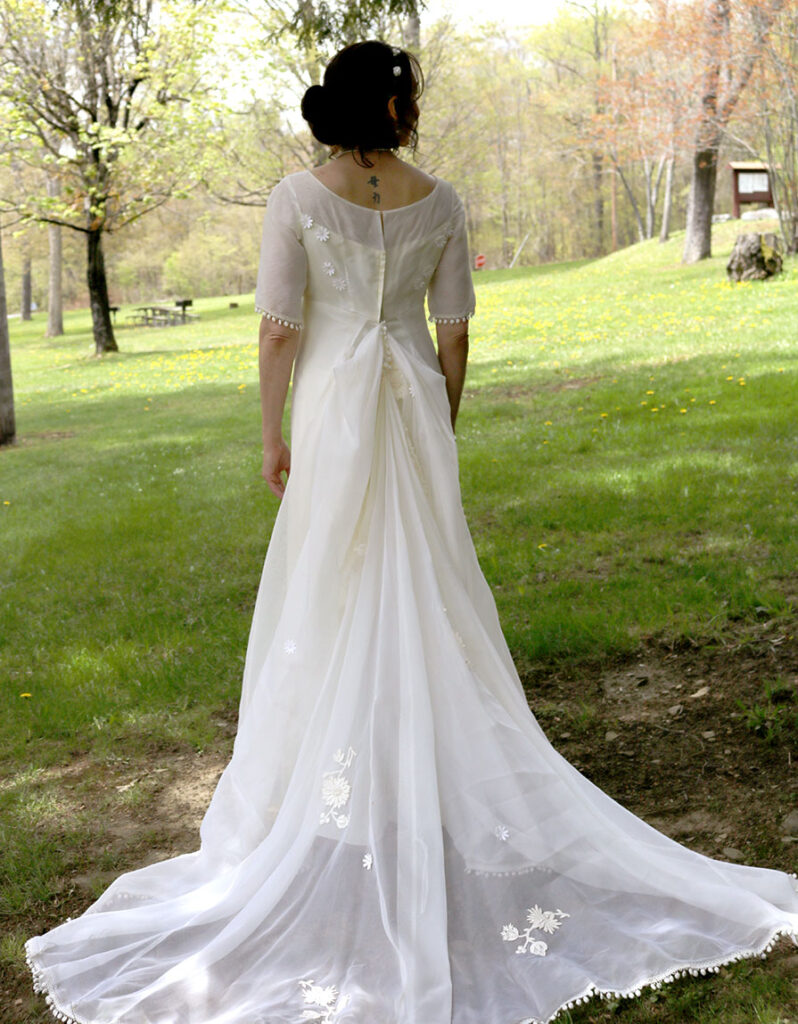 A woman stands in a grassy park, wearing an elegant white wedding dress with a long, flowing train adorned with floral embroidery