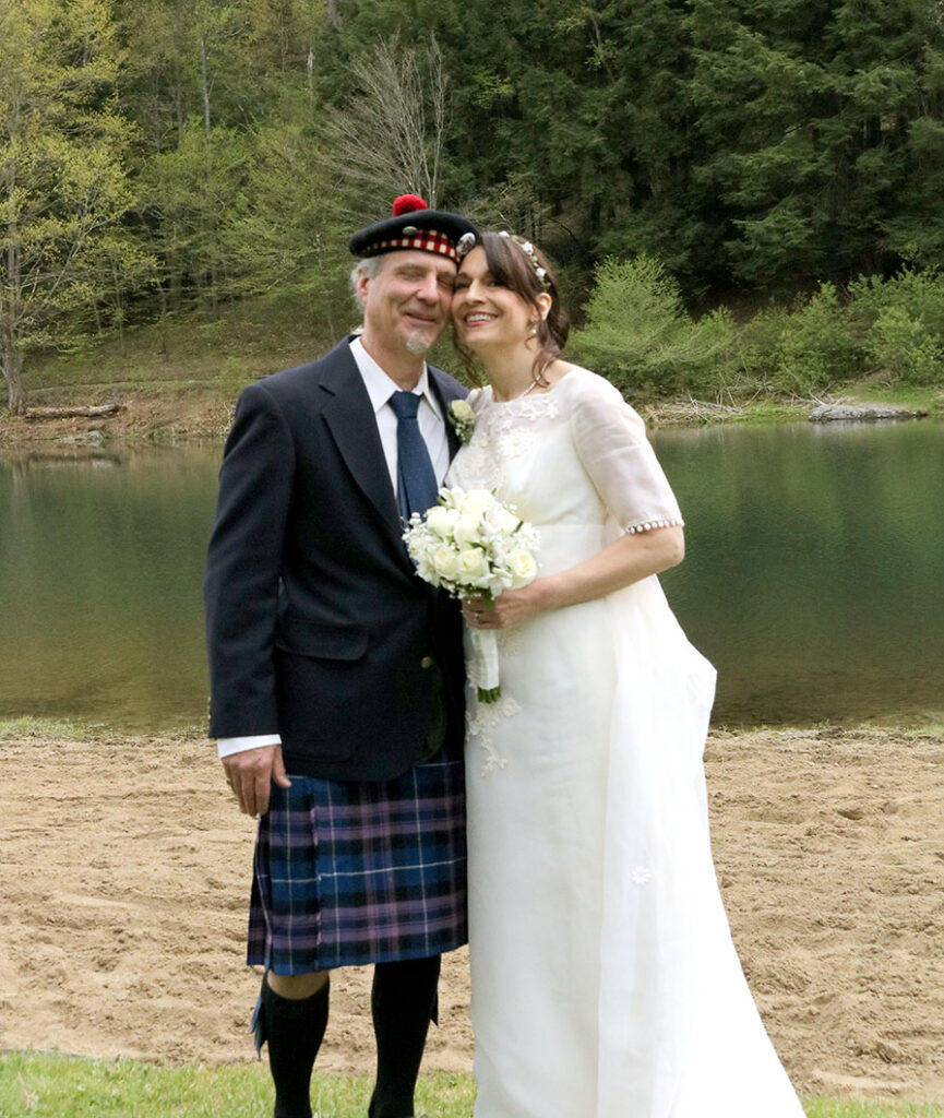 A bride and groom pose happily by a serene lake, surrounded by lush greenery