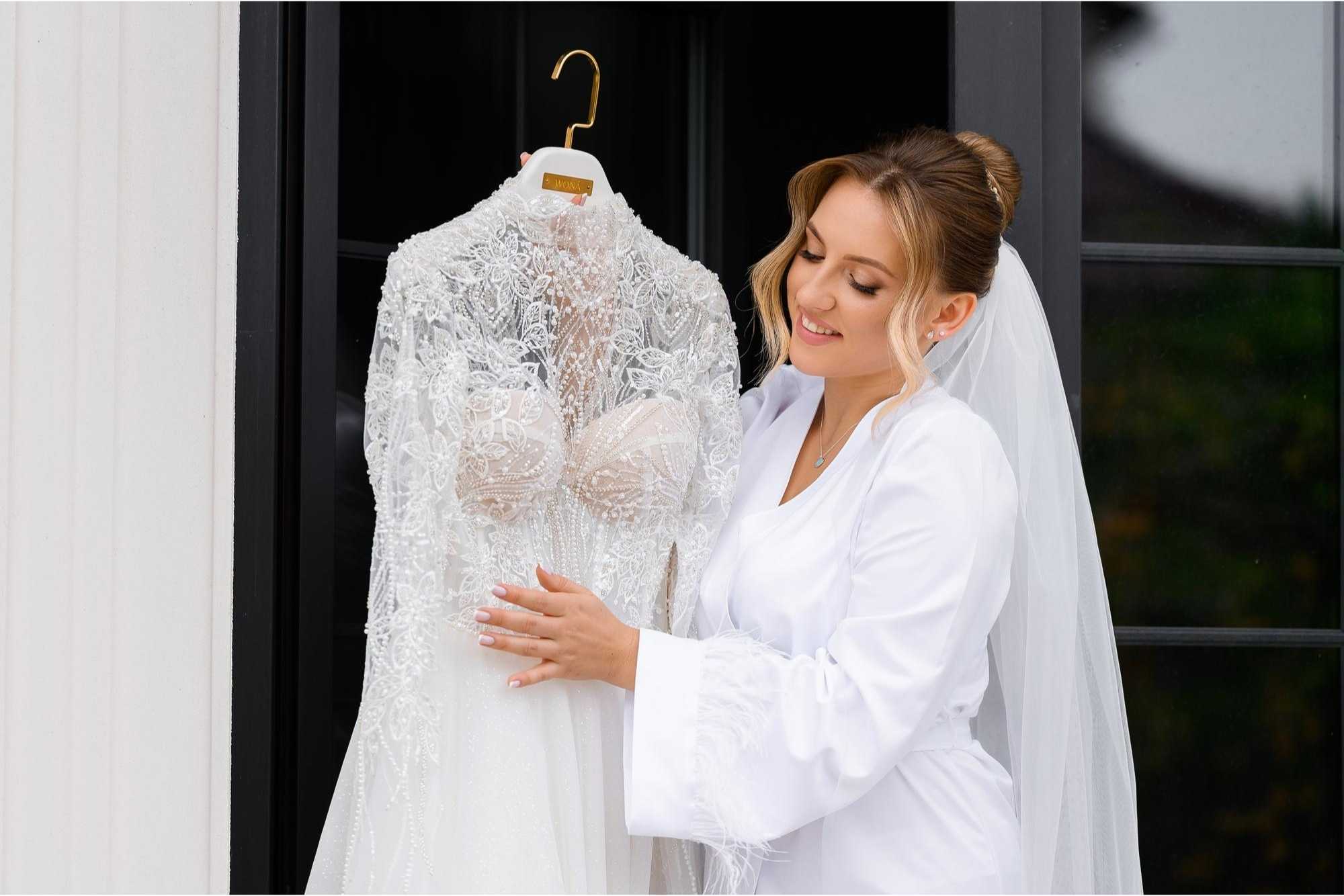 A smiling woman in a white robe and veil admires an intricately detailed wedding dress with lace and beadwork
