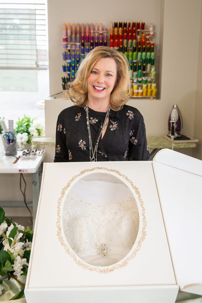 A woman with blonde hair and a floral blouse smiles while standing behind a white box containing a lace wedding dress