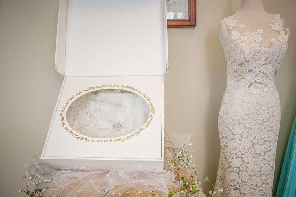A white wedding dress with lace details is neatly folded inside an open box with a decorative gold border