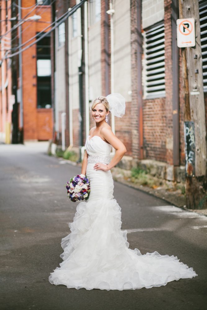 A bride in a flowing white wedding dress poses confidently in an urban alley