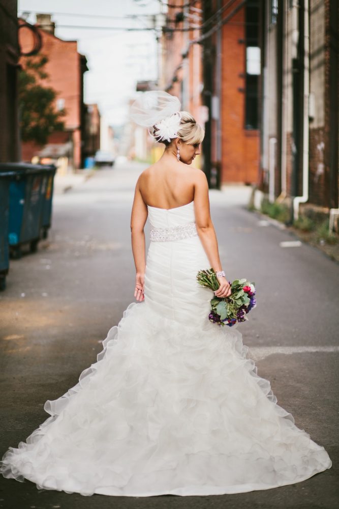 A bride in a strapless white wedding gown stands in an alleyway, holding a bouquet of colorful flowers