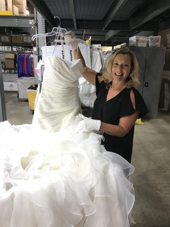 A woman wearing gloves holds up a white wedding dress with a ruffled skirt in a warehouse setting