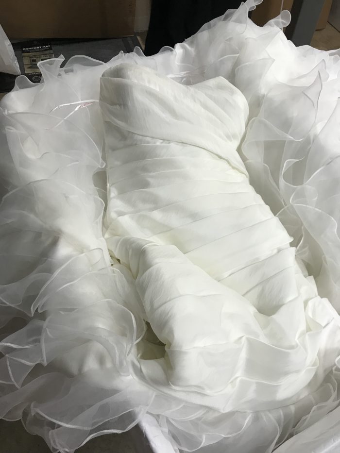 A woman wearing gloves holds up a white wedding dress with a ruffled skirt in a warehouse setting