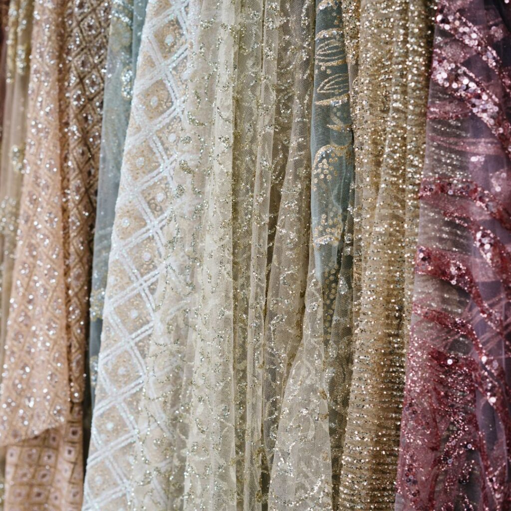 Delicate couture fabrics hanging in a row, featuring intricate beading, sequins, and embroidered patterns in soft neutral and pastel tones.